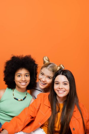 positive blonde Teenager-Mädchen, die in die Kamera in der Nähe von stilvollen multiethnischen Freundinnen mit kühnem Make-up schaut, während sie isoliert auf orangefarbenen, coolen und selbstbewussten multikulturellen Teenager-Mädchen steht, verschiedene Rassen 