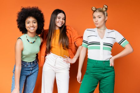 Foto de Adolescentes alegres y multiétnicos con maquillaje audaz posando en trajes de moda y mirando a la cámara juntos sobre fondo naranja, chicas adolescentes multiculturales frescas y seguras - Imagen libre de derechos