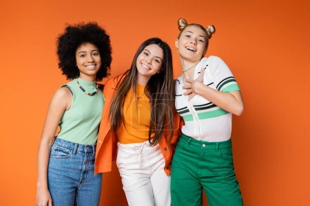 Lächelnder blonder Teenager in lässigem Outfit umarmt gemischtrassige Freundinnen mit kühnem Make-up und blickt in die Kamera auf orangefarbenem Hintergrund, trendige Outfits und modisch zukunftsweisende Blicke