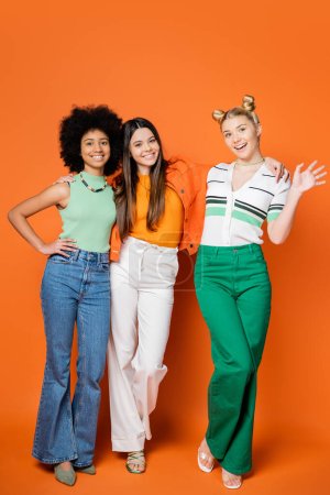 Lächelndes und stilvolles blondes Teenie-Mädchen, das in die Kamera winkt, während es neben multiethnischen Freundinnen steht, mit hellem Make-up auf orangefarbenem Hintergrund, trendigen Outfits und modisch zukunftsweisenden Blicken