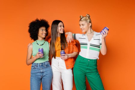 Positive blonde teenager in stylish outfit holding drink in tin can and looking at multiethnic girlfriends in casual outfits on orange background, trendy outfits and fashion-forward looks