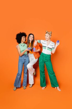Volle Länge positiver und multiethnischer Teenager-Freundinnen in stilvollen Outfits, die Kanner-Drinks halten und auf orangefarbenem Hintergrund posieren, trendige Outfits und modisch nach vorne gerichtete Blicke