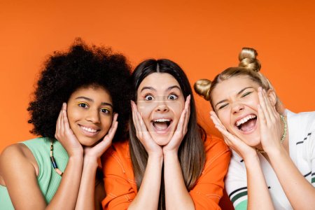 Foto de Emocionadas y alegres novias adolescentes multiétnicas con maquillaje audaz tocando mejillas y mirando a la cámara juntos sobre fondo naranja, trajes de moda y looks de moda, diversas razas - Imagen libre de derechos