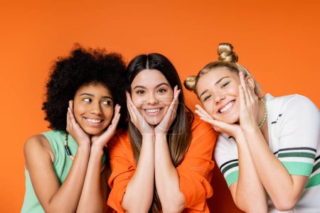 Portrait of positive and interracial teenage girlfriends with bold makeup and casual clothes touching cheeks and standing on orange background, trendy outfits and fashion-forward looks