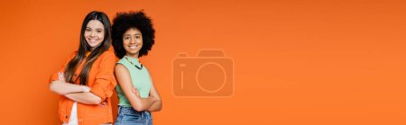Smiling and confident multiethnic teen girlfriends with bold makeup crossing arms and standing back to back isolated on orange, teen fashionistas with impeccable style concept, banner with copy space