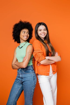Lächelnde afrikanisch-amerikanische Teenager-Mädchen mit kühnem Make-up, die Arme verschränkt und Rücken an Rücken mit stilvoller Freundin auf orangefarbenem Hintergrund, Teen-Fashionistas mit tadellosem Stil-Konzept