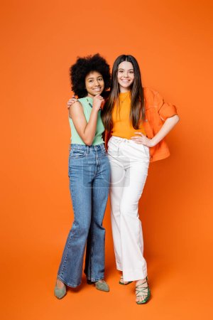 Full length of joyful brunette teen girl in stylish outfit hugging african american girlfriend with bright makeup on orange background, teen fashionistas with impeccable style concept