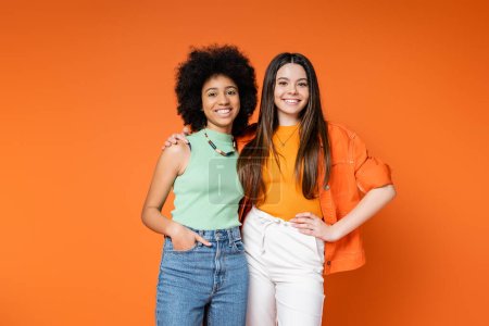 Positive and multiethnic girlfriends with bold makeup and trendy outfits hugging and looking at camera while posing isolated on orange, teen fashionistas with impeccable style concept