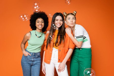Positive and interracial girlfriends in stylish outfits hugging and posing near soap bubbles and standing on orange background, teen fashionistas with impeccable style concept, diverse races 