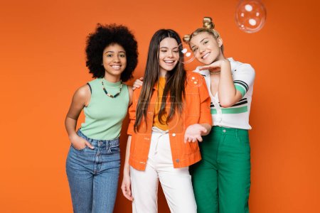 Joyful and multiethnic teen girlfriends in trendy casual outfits posing and standing near soap bubbles on orange background, teen fashionistas with impeccable style concept