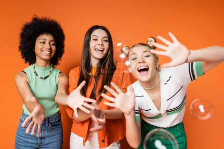 Photo for Excited and cheerful multiethnic teen girls with bold makeup looking at soap bubbles while posing and standing on orange background, teen fashionistas with impeccable style concept - Royalty Free Image