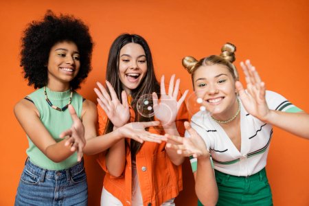 Photo for Cheerful and multiethnic teen girlfriends in stylish casual clothes looking at soap bubbles while standing on orange background, teen fashionistas with impeccable style concept - Royalty Free Image