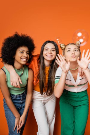 Cheerful brunette teenager hugging stylish multiethnic girlfriends and standing near soap bubbles on orange background, multiethnic teen fashionistas with impeccable style concept
