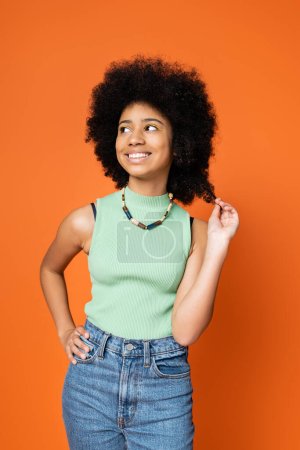 Stylish african american teenage girl in casual outfit and necklace touching hair, holding hand on hip and looking away isolated on orange, trendy teenage girl expressing individuality