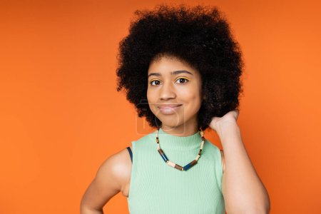 Portrait of smiling and stylish african american teenager with bold makeup wearing necklace and touching hair while looking at camera isolated on orange, trendy teenage girl expressing individuality