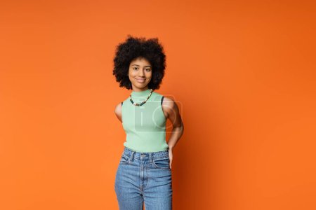Lächelndes und stilvolles afrikanisch-amerikanisches Teenager-Mädchen in lässiger Kleidung mit Halskette und fettem Make-up posiert und schaut isoliert in die Kamera auf orangefarbenem, trendigem Teenager-Mädchen, das Individualität ausdrückt