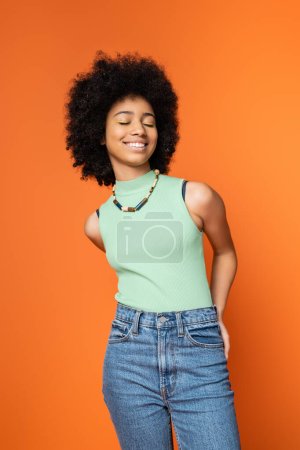 Freudiges und stilvolles afrikanisch-amerikanisches Teenager-Mädchen mit Halskette und fettem Make-up, das die Augen schließt, während es isoliert auf orangefarbenen, trendigen Teenager-Mädchen steht, die Individualität ausdrücken