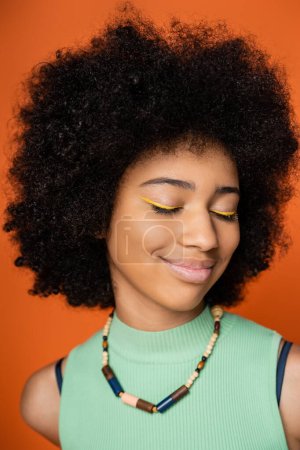 Porträt eines fröhlichen und jugendlichen afrikanisch-amerikanischen Mädchens mit kühnem Make-up und stilvoller Halskette, die die Augen schließt, während sie isoliert auf einem orangefarbenen, trendigen Teenager-Mädchen steht, das Individualität ausdrückt