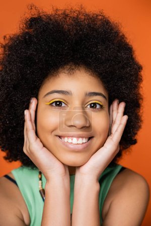 Portrait of positive and stylish african american teenager with bold makeup touching face and looking at camera while posing isolated on orange, trendy teenage girl expressing individuality 