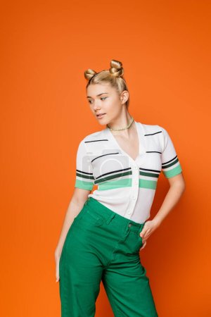 Trendy and confident blonde teenage girl with bold makeup posing in casual outfit and looking away while standing on orange background, trendy teenage girl expressing individuality 