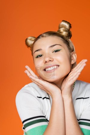 Portrait of positive blonde teenager with hairstyle and bold makeup holding hands near face while posing isolated on orange, trendy teenage girl expressing individuality, gen z
