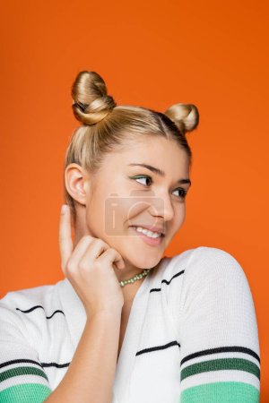 Portrait of smiling and trendy blonde teen girl with hairstyle and bold makeup pointing with finger and looking away while standing isolated on orange, trendy teenage girl expressing individuality