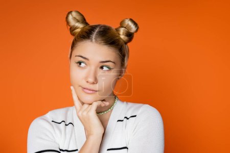 Porträt eines nachdenklichen und stilvollen Teenager-Mädchens mit Frisur und kühnem Make-up, das wegschaut, während es in lässiger Kleidung isoliert auf orangefarbenen, trendigen Teenager-Mädchen posiert, die Individualität ausdrücken