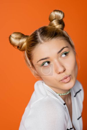 Porträt eines verträumten und trendigen blonden Teenie-Mädchens mit Frisur und kühnem Make-up, das wegschaut, während es in lässiger Kleidung posiert und isoliert auf orangefarbener, modischer und trendiger Kleidung steht