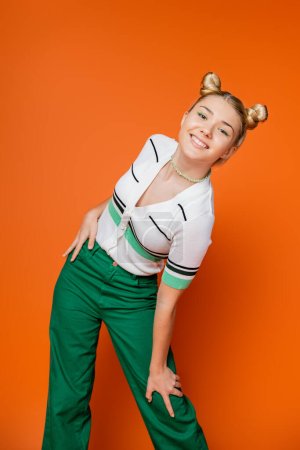 Positive and fashionable teenage girl with hairstyle and bold makeup posing in casual outfit and looking at camera while standing on orange background, fashionable and trendy clothes