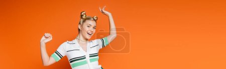 Aufgeregter und fröhlicher blonder Teenager mit Frisur und kühnem Make-up posiert in lässiger Kleidung und blickt in die Kamera, während er isoliert auf orangefarbener, modischer und trendiger Kleidung steht, Banner 
