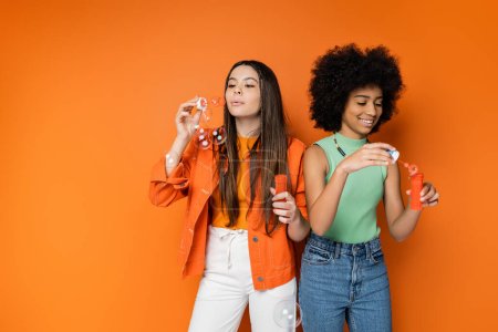 Stilvolle brünette Teenager-Mädchen weht Seifenblasen in der Nähe fröhliche afrikanisch-amerikanische Freundin mit kühnem Make-up, während sie zusammen auf orangefarbenem Hintergrund, modische und trendige Kleidung