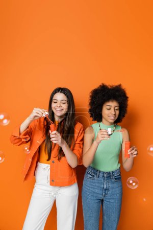 Lächelnde Brünette und afrikanisch-amerikanische Teenager-Freundinnen mit fettem Make-up in lässigen Outfits, die Seifenblasen halten und auf orangefarbenem Hintergrund stehen, modische und trendige Kleidung