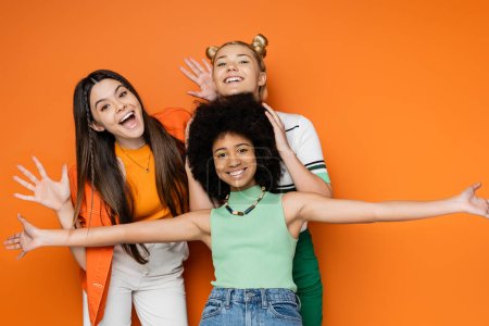 Cheerful and multiethnic teenage girls with colorful makeup posing together in casual outfits and looking at camera while standing on orange background, fashionable and trendy clothes
