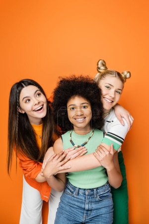 Adolescents positifs avec un maquillage coloré et tenues décontractées embrassant petite amie afro-américaine ensemble et posant sur fond orange, vêtements à la mode et à la mode