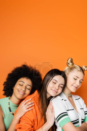 Novias adolescentes multiétnicas positivas y elegantes con maquillaje colorido que usan trajes casuales mientras se abrazan entre sí con ojos cerrados aislados en ropa naranja, de moda y de moda.