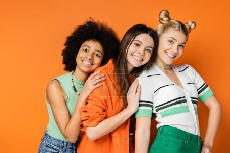 Lebendige multiethnische Teenager mit kühnem Make-up tragen stylische Freizeitkleidung und umarmen ihre blonde Freundin, während sie zusammen auf orangefarbenem Hintergrund stehen, modische und trendige Kleidung