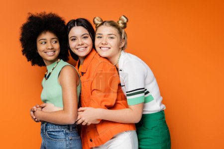 Positiver blonder Teenager mit kühnem Make-up umarmt stilvolle multiethnische Freundinnen in lässigen Outfits und blickt in die Kamera auf orangefarbenem Hintergrund, modische und trendige Kleidung