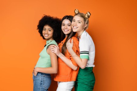 Modernas y sonrientes novias adolescentes multiétnicas con maquillaje audaz que usan atuendos casuales mientras posan y miran a la cámara en un fondo naranja, poses elegantes y confiadas