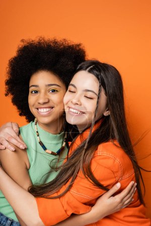 Lächelnd und stilvoll afrikanisch-amerikanische Teenager-Mädchen mit kühnem Make-up umarmt brünette Freundin und blickt in die Kamera, während sie isoliert auf orangefarbenen, stilvollen und selbstbewussten Posen steht