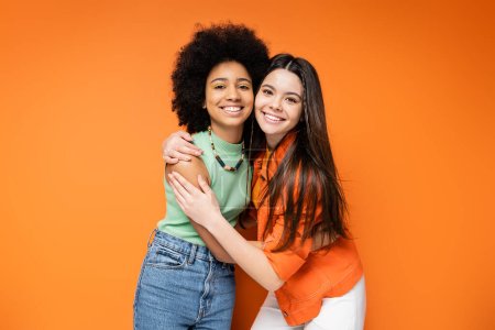 Interracial and stylish teenage girlfriends with bold makeup wearing casual outfits and hugging each other while posing on orange background, stylish and confident poses