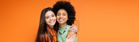 Lächelnder brünetter Teenager in lässiger Kleidung umarmt afrikanisch-amerikanische Freundin mit kühnem Make-up und blickt isoliert in die Kamera auf orangefarbenen, stilvollen und selbstbewussten Posen, Banner 