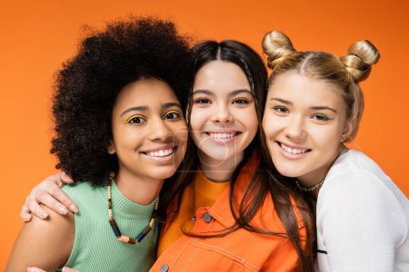 Porträt eines lächelnden Teenagers mit buntem Make-up, der multiethnische Freundinnen umarmt und gemeinsam in die Kamera schaut, während er isoliert auf orangefarbenen, stilvollen und selbstbewussten Posen posiert