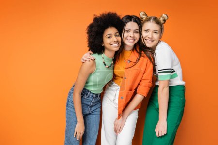 Chicas adolescentes animadas y multiétnicas con maquillaje colorido y ropa casual abrazando y posando juntos mientras están de pie sobre fondo naranja, poses elegantes y confiadas