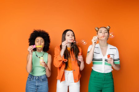 Multiethnic teenage girlfriends with bold makeup and casual outfits blowing soap bubbles and spending time together while standing on orange background, trendy and stylish hairstyles