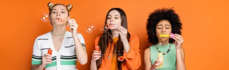 Interracial teenage girlfriends with colorful makeup wearing casual clothes while blowing soap bubbles and spending time on orange background, trendy and stylish hairstyles, banner 
