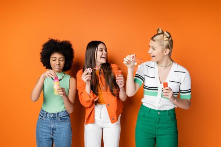 Foto de Chicas adolescentes positivas y de moda con maquillaje colorido que usan trajes casuales y la celebración de burbujas de jabón cerca de la novia afroamericana sobre fondo naranja, peinados de moda y con estilo - Imagen libre de derechos