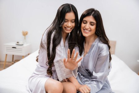 Photo for Happy asian woman and her friend sitting on bed, showing engagement ring, happiness, bridal party, silk robes, best friends, bride with her bridesmaid, brunette hair, cultural diversity, multiracial - Royalty Free Image