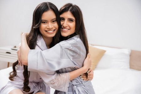 Photo for Happy asian woman and her friend sitting on bed, hugging each other, bridal party, silk robes, best friends, bride with her bridesmaid, brunette hair, cultural diversity, multiracial, pre-wedding - Royalty Free Image