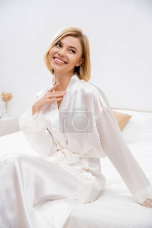fröhliche Braut mit blonden Haaren im weißen Seidenmantel auf dem Bett sitzend, wegschauend, lächelnd, Hochzeitsplanung, junge Frau, schön, aufregend, feminin, selig 