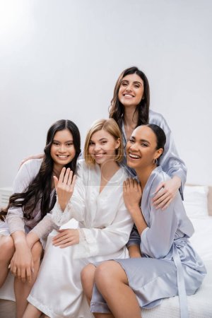 vier Frauen, Brautparty, glückliche blonde Braut zeigt Verlobungsring in der Nähe ihrer interrassischen Brautjungfern, auf dem Bett sitzen, Glück, Seidenroben, brünett und blond, beste Freunde, Vielfalt